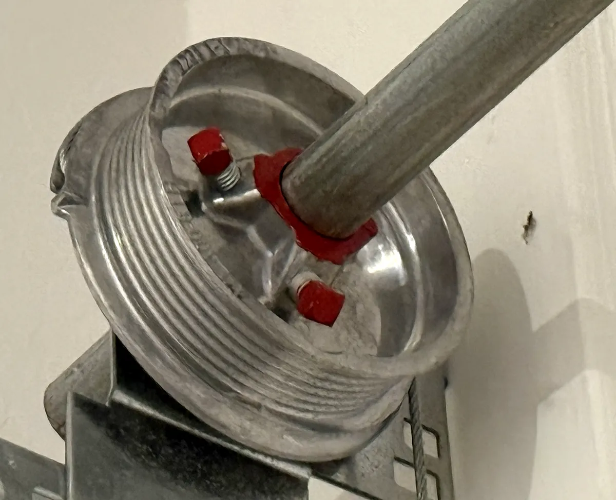 A garage door cable drum on the spring bar above a West Valley garage door, with grooves for the lift cable, red set screws and the cable running down past the end bracket