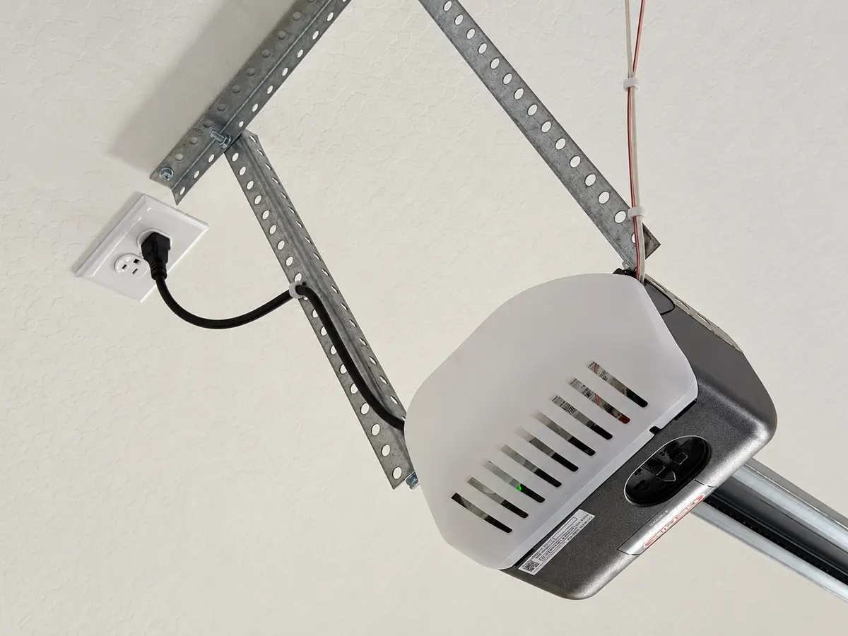 A ceiling-mounted garage door opener with its light cover, power cord and the rail running toward the door
